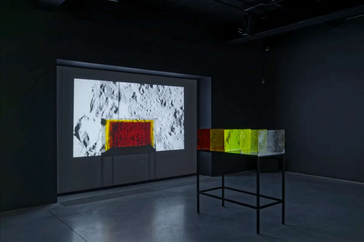 A dark gallery with a black table in the middle of the floor with colored glass plates on top of it which are projected onto a screen.