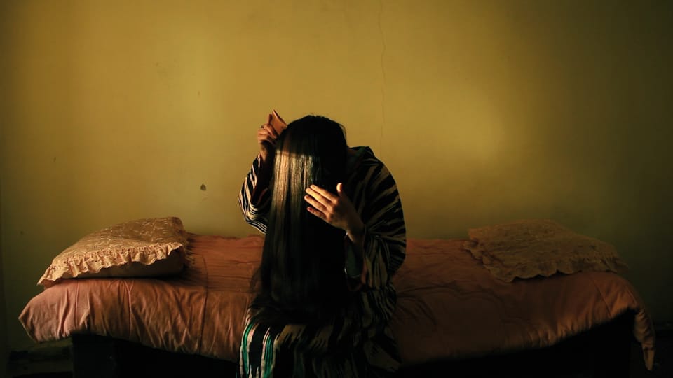 A woman sitting on a bed, combing her long black hair forward over her face.