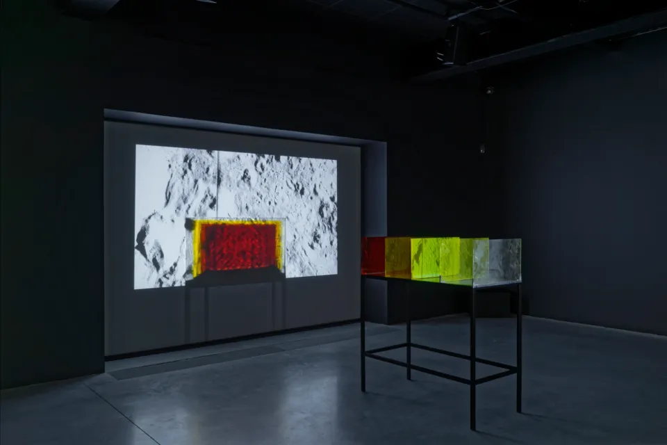 A dark gallery with a black table in the middle of the floor with colored glass plates on top of it which are projected onto a screen.